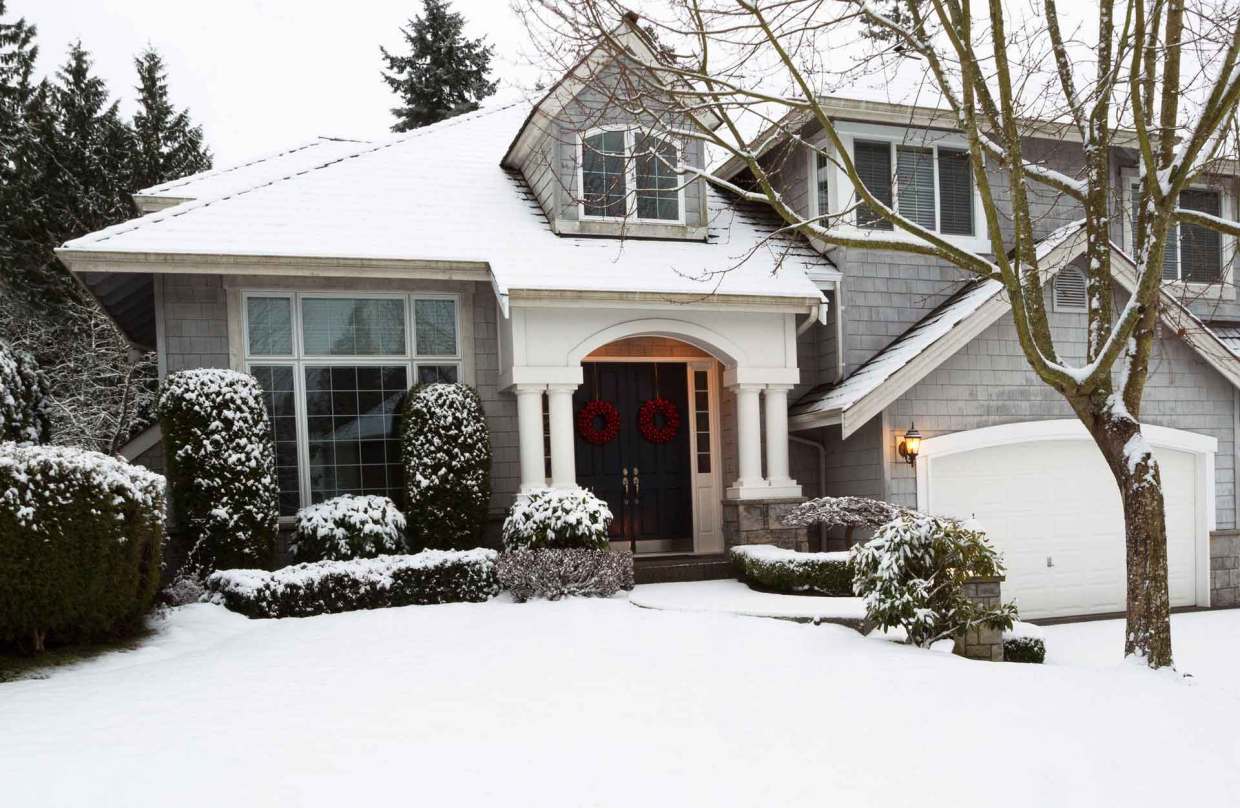 The exterior of a gray house with the ground, bushes, and trees covered in snow.