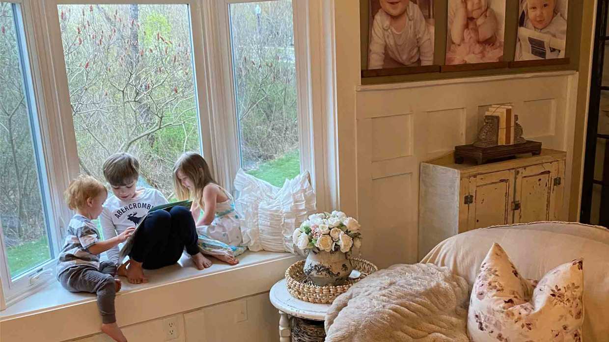 Three children sit in a bay window sill reading a book together.