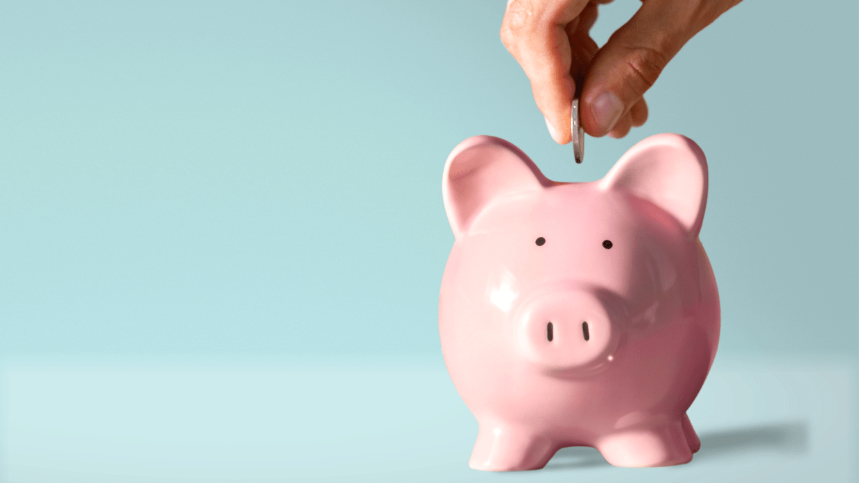 A hand puts a coin in a pink piggy bank