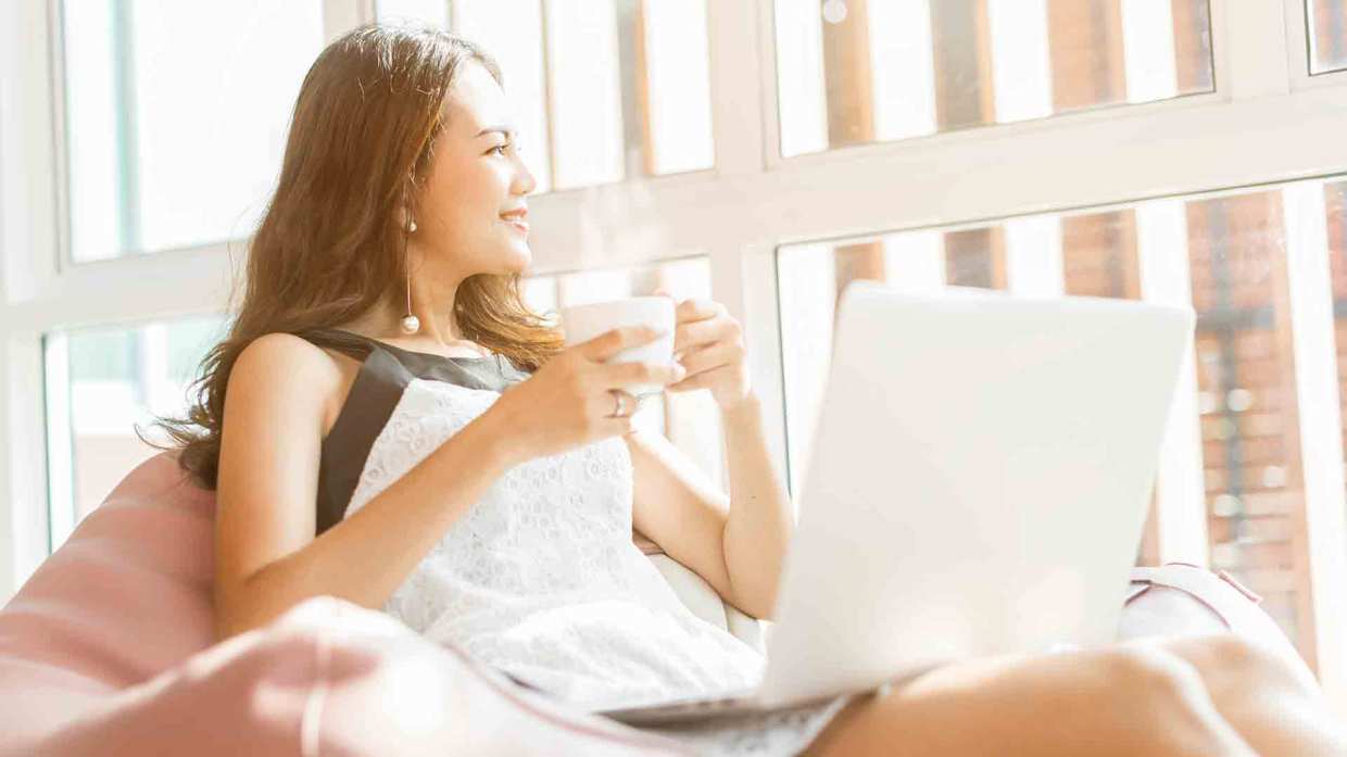 A woman relaxes in front of a sunny window with a coffee cup and a laptop on her lap.