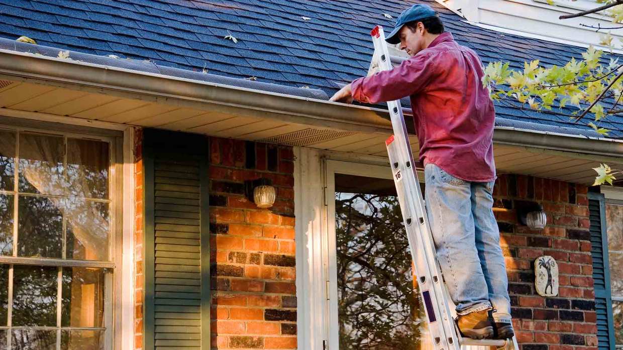 A man stands on a latter resting on the side of his house to inspect and clean out his gutters.