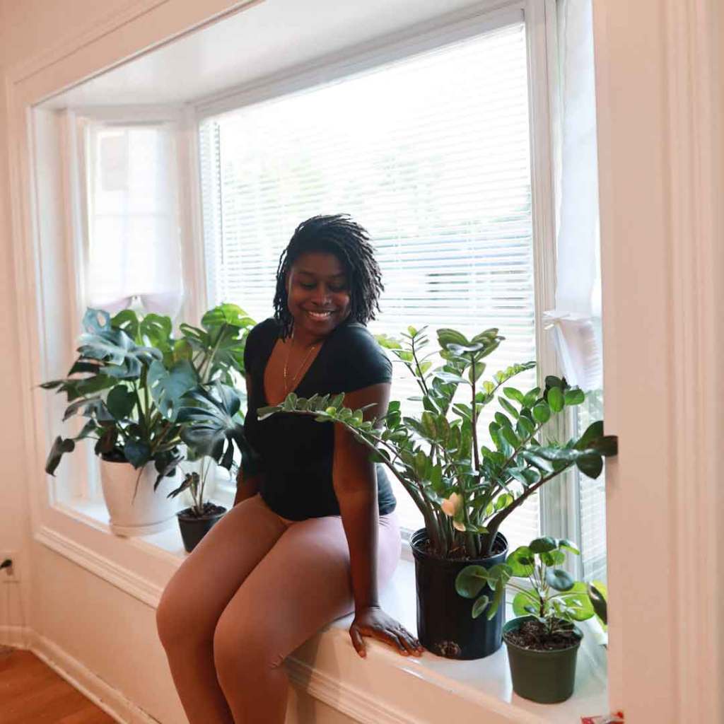 A woman sits happily in her bay window installed by Universal Windows Direct surrounded by houseplants.