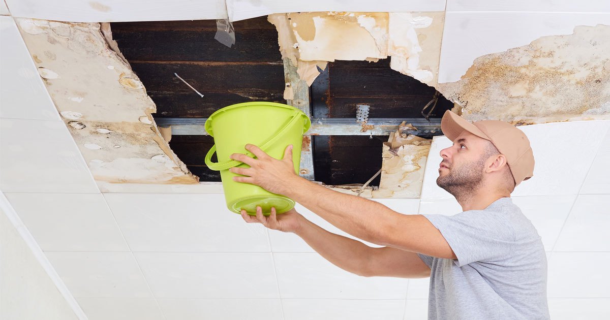 A man holds a bucket to a torn and leaking ceiling.