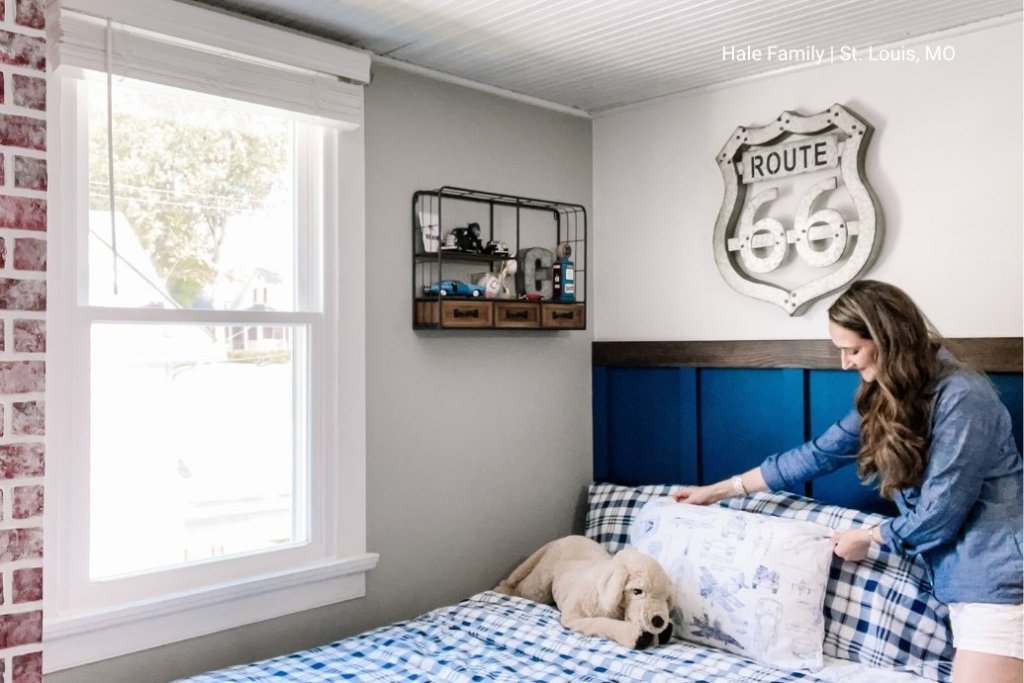 A woman fluffs a pillow on a bed with a bright double hung window nearby.