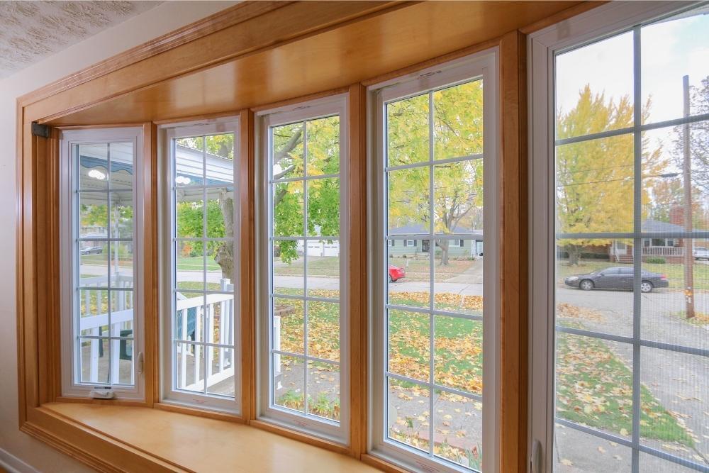 A view through a bow window from the inside, looking out to a front yard with fall leaves.