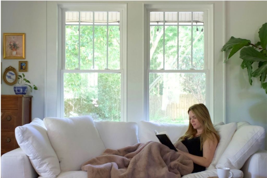 A woman reads a book under a blanket on her couch in front of two bright windows.