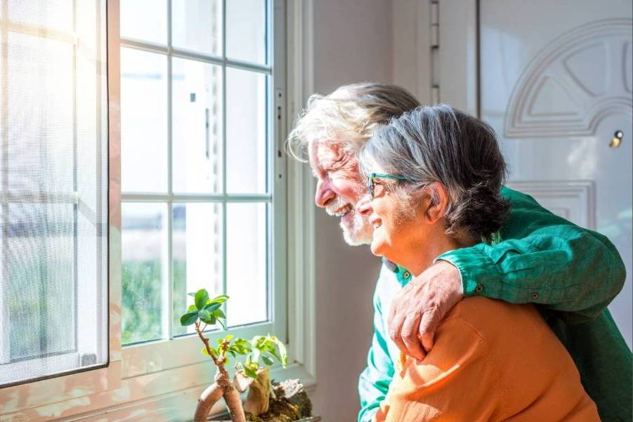 A happy senior couple looks out of the sunny window from inside their home.