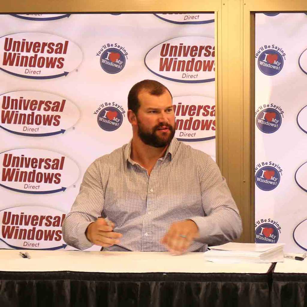 Joe Thomas seated at a signing table in front of a Universal Windows Direct backdrop